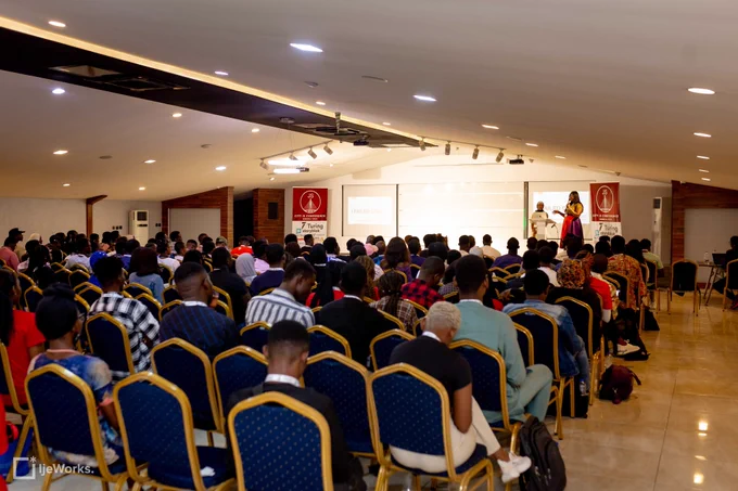 The view from back of the conference hall showing all attendees at the event including the host and speaker on stage.