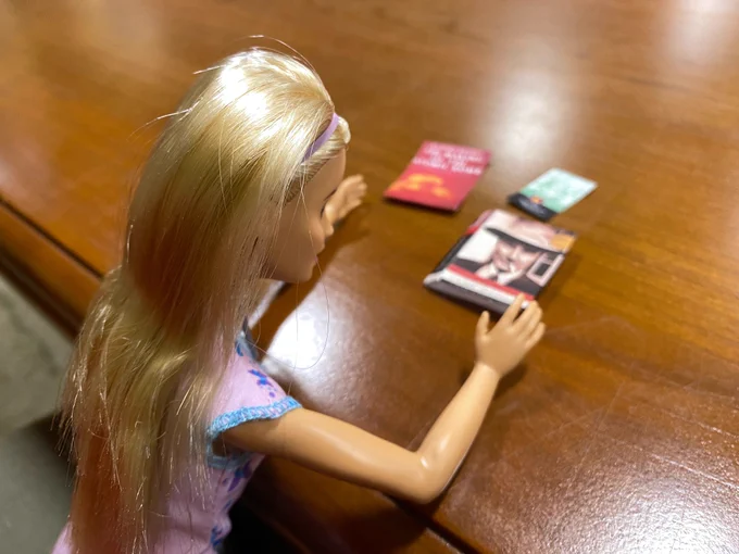 Photo of nurse Barbie doll at table, with hands near (photos of) 3 books about the atomic bomb: The Making of the Atomic Bomb (1986) by Richard Rhodes (red cover) & The Manhattan Project: Big Science and the Atom Bomb (2003) by Jeff Hughes (green cover) & American Prometheus: The Triumph and Tragedy of J. Robert Oppenheimer (2005) by Kai Bird and Martin J. Sherwin (photo of Oppenheimer on cover).