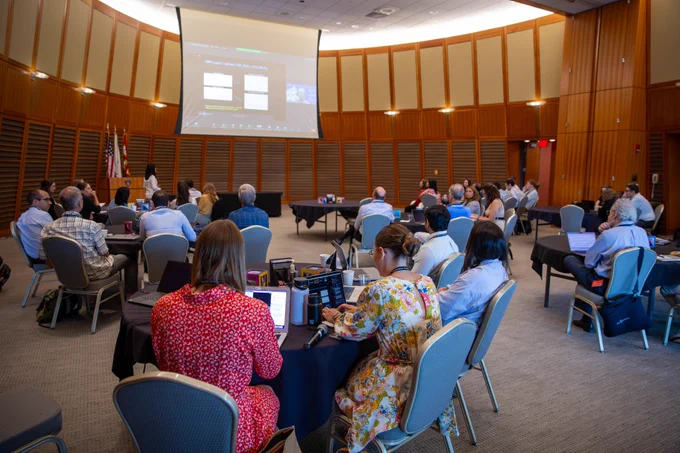 Large room of people sitting at tables watching someone present with slides on a large screen