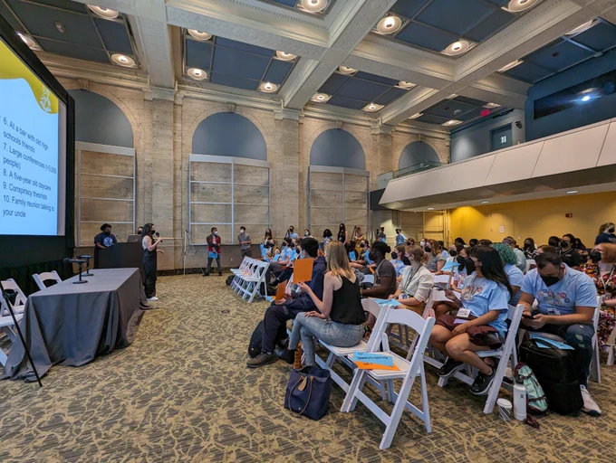 Conference venue with Ashley Kim presenting at the front of the room and attendees sitting in the audience holding AWESOME/JARGON signs