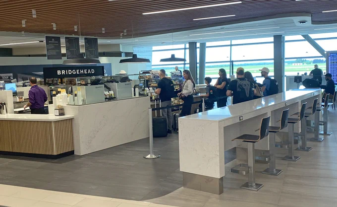 People lined up at a counter. Sign with ‘Bridgehead’ on it above counter. Counter-height stools along a white marble counter.