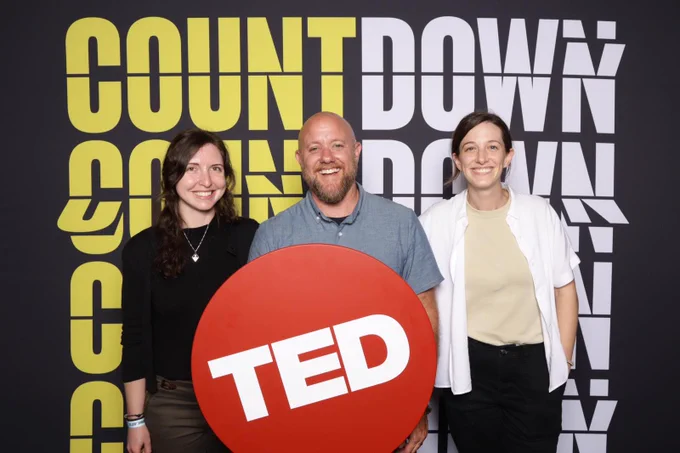 Man and two women standing in front of a black backdrop with the word Count Down written in bold yellow and white letters over and over again. The man is holding a round red sign that says Ted on it with white text. 