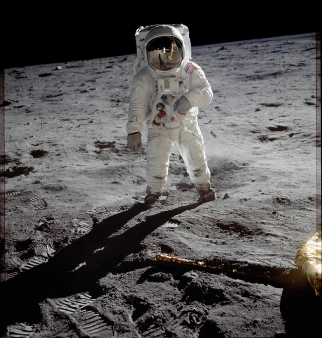 Photo of astronaut Buzz Aldrin standing on the surface of the Moon in his white spacesuit. Reflected in his visor is Neil Armstrong, who took the photo. Bootprints on the fine, gray lunar soil are visible in the foreground. A portion of the lunar lander's landing leg is in the bottom right of the image.