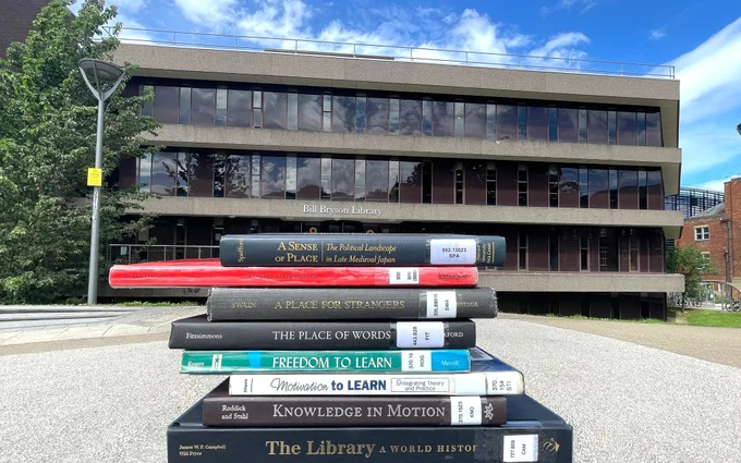 A series of books stacked in a pile with the book spines facing towards the camera. Library Square and Bill Bryson Library are in the background.