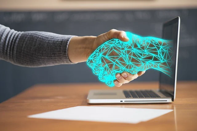 A human-like hand made of bright blue geometric lines reaches from out of a laptop screen to grasp a person's hand in a handshake