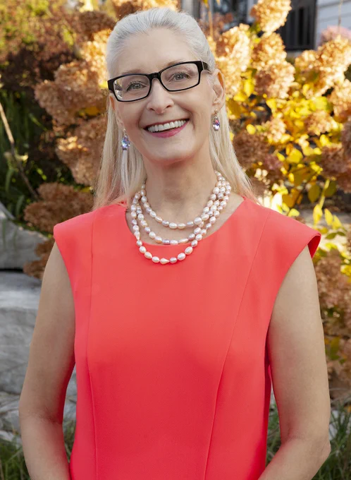 author photo: Lisa de Nikolits. She is a white woman with long blonde hair. She wears eyeglasses, a coral blouse, a three-string necklace, and a big smile. She stands outside in front of bright yellow and gold flowers/greenery.