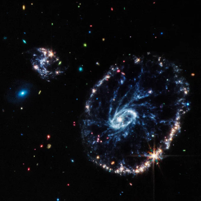 Webb's mid-infrared image of the Cartwheel Galaxy. It shows a blue ring studded with bright points, connected to a ghostly pale blue core by faint blue threads of dust resembling wheel spokes. Unlike the composite image, there appears to be no material between the "spokes." At the upper left are two small companion galaxies. The one on the top is shaped like a backwards S and marked by bright points amidst blue threads. The one below it looks like small glowing blue oval. The background is dark and filled with colorful dots in green, red, blue and more.