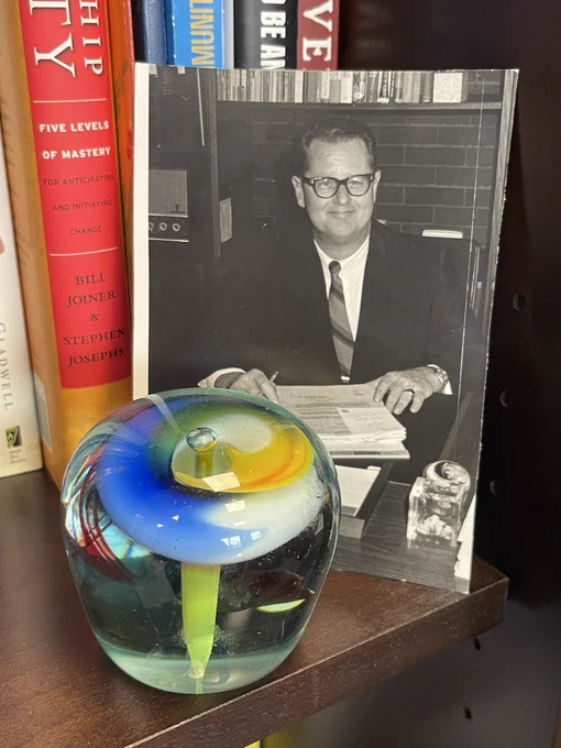 Black and whit snapshot of Kenneth W. Robinson seated at a desk. In the foreground is a colorful glass paperweight; this paperweight is one of the items on the desk in the photo.
