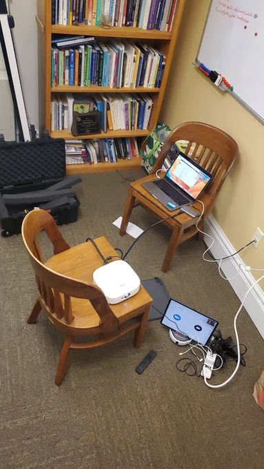 Pic of office with 2 chairs, an open travel case, a laptop, projector and SWiVL robot and lots of cords. With overfull bookshelf in background. 