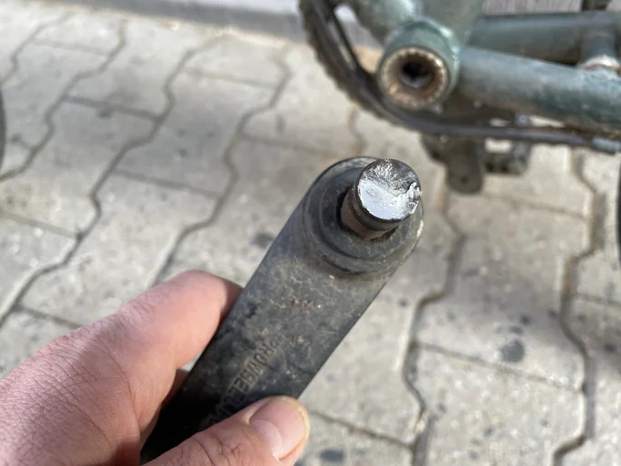 Eine Hand hält eine Fahrradkurbel ins Bild, die Achse in der Mitte ist durchgebrochen, man sieht eine aluminiumschimmernde Bruchkante. Im Hintergrund das Fahrrad, von dem die Kurbel abgebrochen ist
