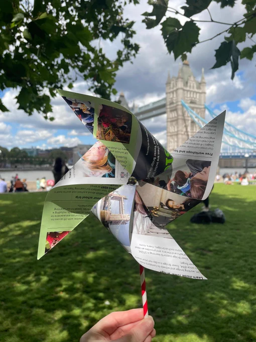 A paper windmill held up in front of Tower Bridge framed by grass and tree branches.