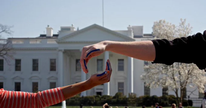 two hands with a blue streak on the side come together to make a zero in front of the White House