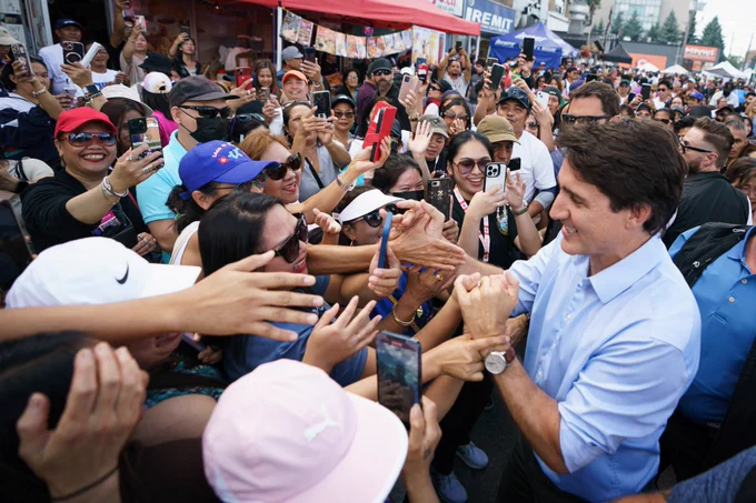 Prime Minister Justin Trudeau is shaking hands with a person. A large crowd of people is standing in front of him and beside him.