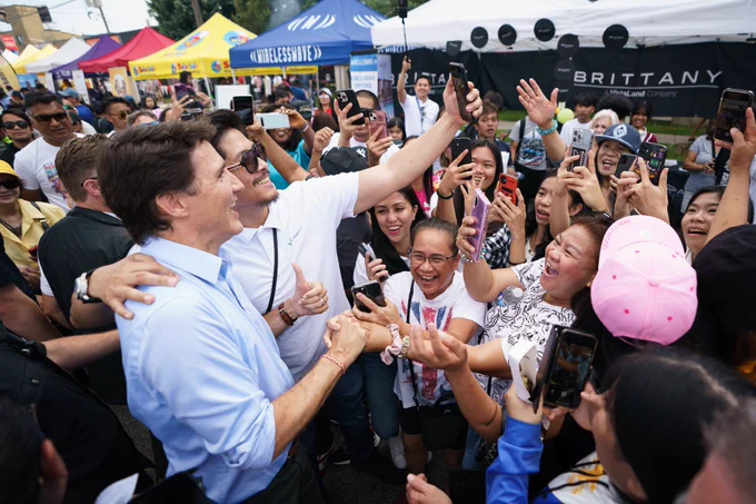 Prime Minister Justin Trudeau is standing and posing for a picture with a man. The man is holding his cellphone in the air and taking a selfie. A large crowd of people is standing in front of him.