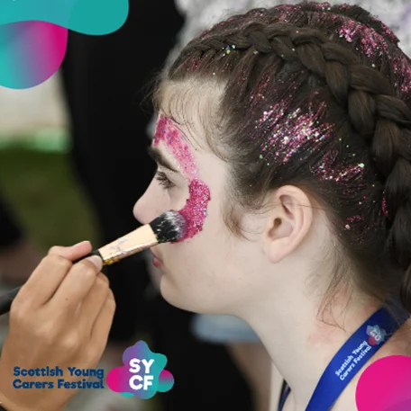 One girl having glitter painted on her face. Her hair is also braided with glitter. There are pink, blue and purple blob shapes at the borders of the image. 