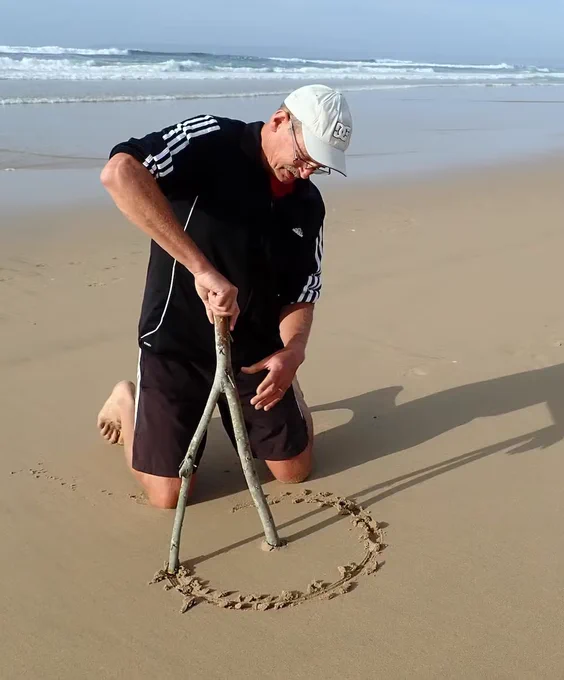 Man in black attire on his knees on cream sand, demonstrating how one of the ammoglyphs was probably made. He is holding a stick with two branches. One branch is anchored in the sand, allowing rotation of the other brand to draw a circle in the sand.