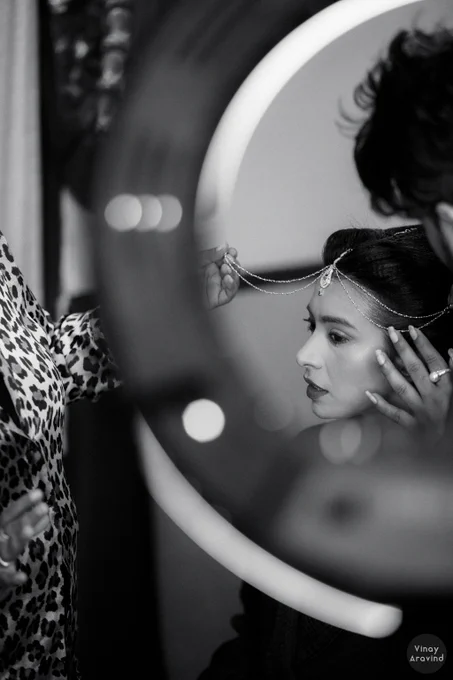 Black and white image of a bride being dressed up. FBride's face is visible on the right edge of the frame shot through an out of focus ring light in the foreground. A make-up artist's head is visible on the top right and a leopard print pattern is visible on the left. 