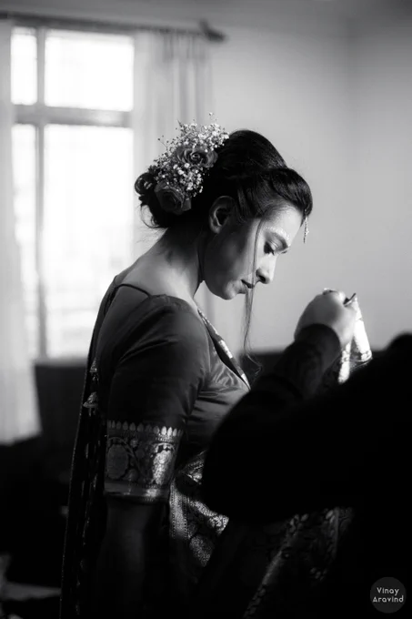 Black and white image of a bride being assisted with her sari. Mid shot of the bride showing her right profile. There's baby's breath in her hair. Gilded border of the blouse is visible. A hand in a black full sleeve is seen holding the sari pallu on the bottom right of the frame. There is a bright out-of-focus window in the background.