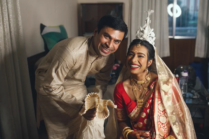 Colour picture of bride and groom laughing at the camera. Bride is wearing a red and  gold sari with traditional Bengali make-up and topor on her head. Groom is wearing a beige kurta and holding the end of his dhuti in his hand. 