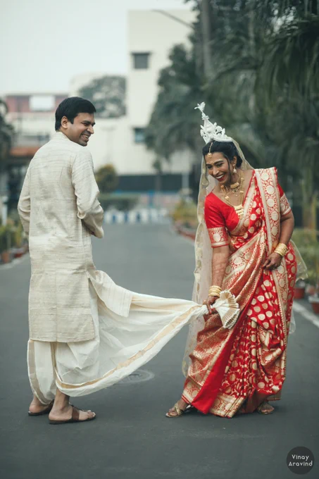 Full length picture of bride and groom on a road. Both are laughing. Groom is turning away from camera. Bride (in red and gold sari) is facing camera,  leaning to her right and pretending to yank the groom's (in beige kurta) dhuti. 