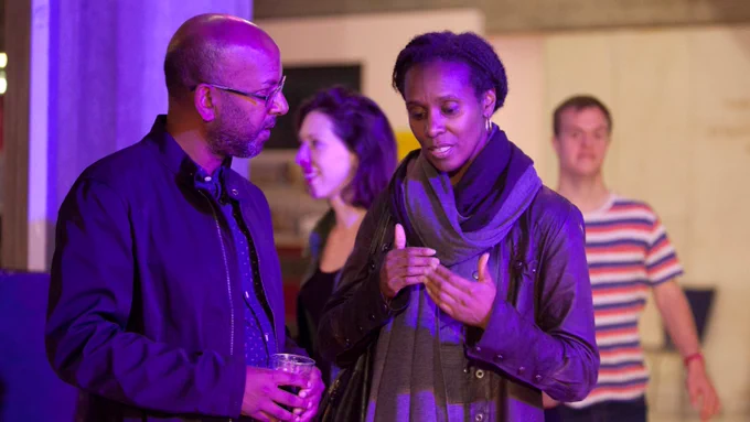 Two people stand speaking to each other at an event. They are lit by a purple light from above. One of them is holding a drink, and the other is moving their hands in an expressive way as they speak.