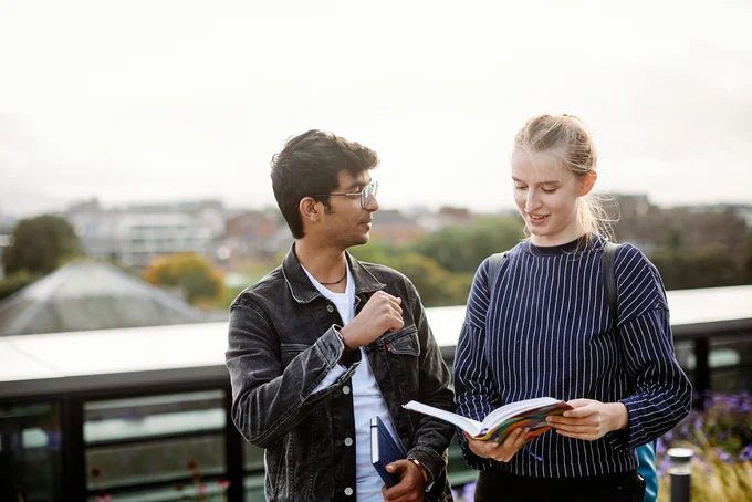 Two international students stand discussing a project they are to work on together. One is holding a book open to show the other student where to find the questions for the project.  