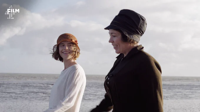 Jessie Buckley, who plays Rose, and Olivia Colman, who plays Edith, walk alongside each other on a beach, smiling and wearing 1920s attire.