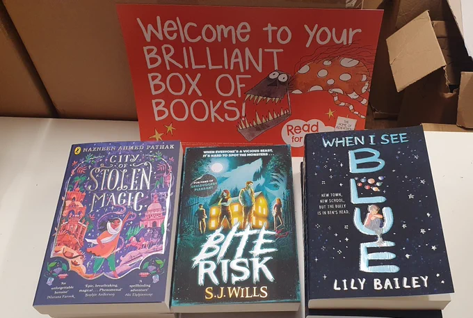3 paperback books on a table, from left to right - City of Stolen Magic, Bite Risk, When I See Blue. Behind the books is an orange poster which says Welcome to Your Brilliant Box of Books 