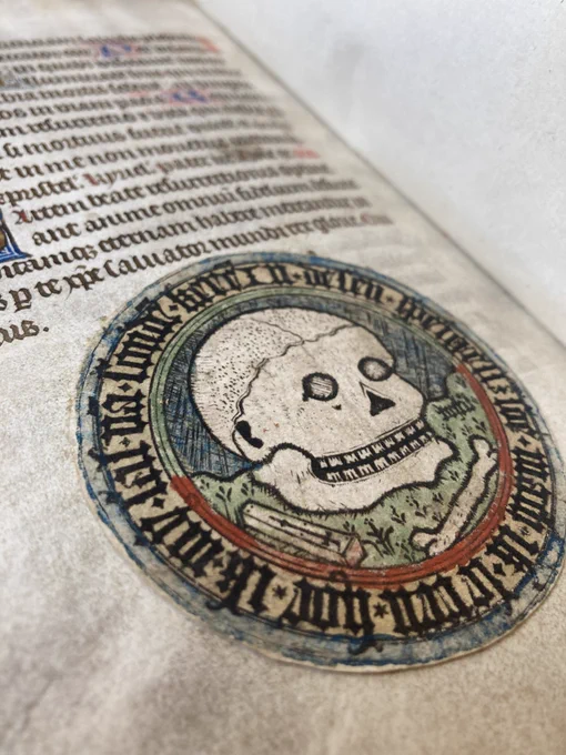 A circular paper emblem of a skull, coffin, and bone attached to f. 167v in TCD MS 103 Book of Hours. 