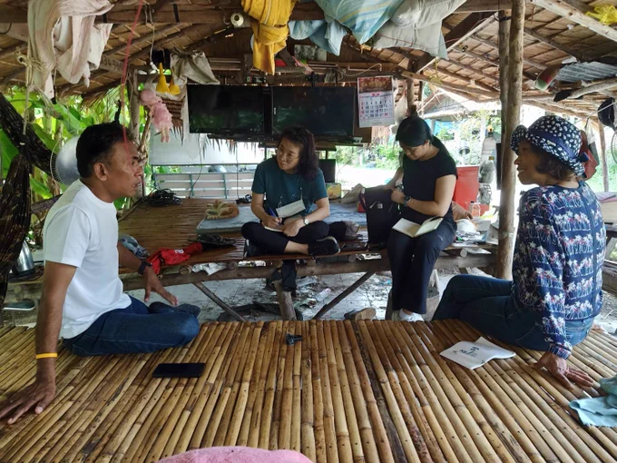 Geography researchers listening to a Thai small-scale fisher under the shade of a roof on the edge of the beach. 