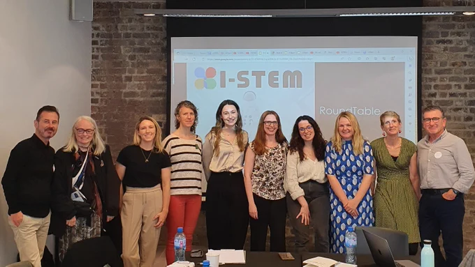 10 people smiling indoors in front of a screen that says "I-STEM".