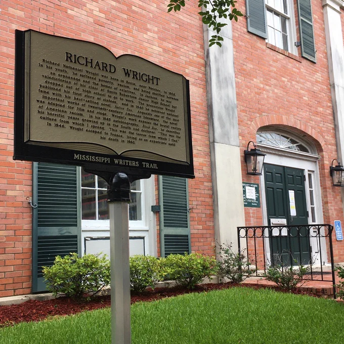 A Mississippi Writers Trail sign talking about Richard Wright sits in front of a large brick building, the Natchez Public Library.