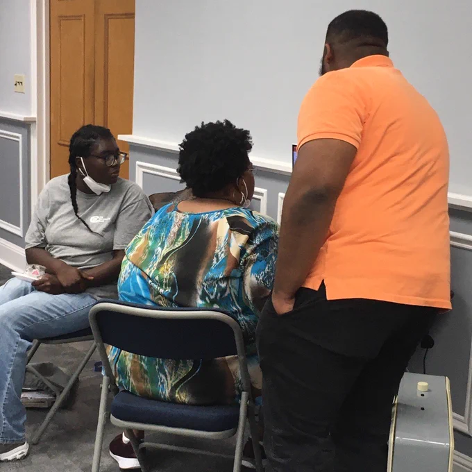 Three Black people, of various ages, look at a computer. We can't see what's on the computer screen, but the two younger people look to be helping an older woman.