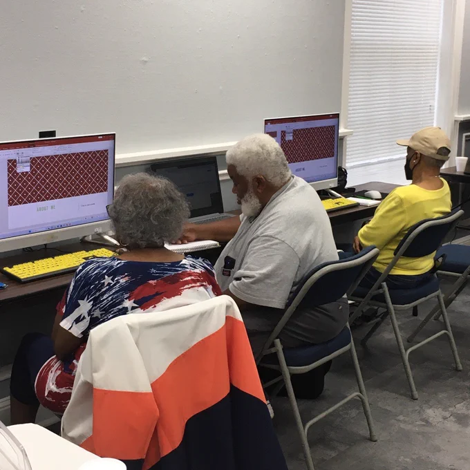 Three Black people sit at two computers. From left to right: a Black woman with gray hair and a patriotic themed shirt, a Black man with white hair, a white beard, and a gray shirt, a Black woman with a tan baseball cap and a yellow shirt. Both computers display the same PowerPoint presentation.