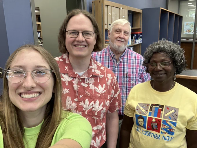 Four people smile at the camera. Left to right: a white woman with glasses and a green shirt; a white man with glasses and a Hawaiian shirt, a white man with white hair, a beard, and a plaid shirt, a Black woman with gray hair, glasses, and a yellow shirt that reads "All Together Now."