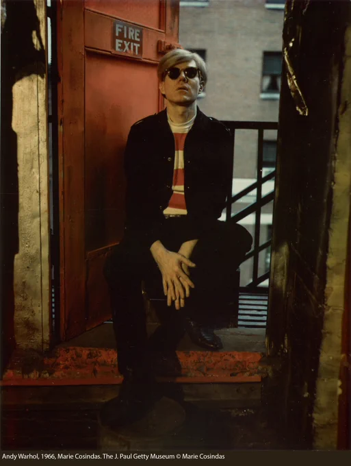 A person with blond hair wearing sunglasses sits in a doorway in front of an open orange-red door with a sign that reads FIRE EXIT.