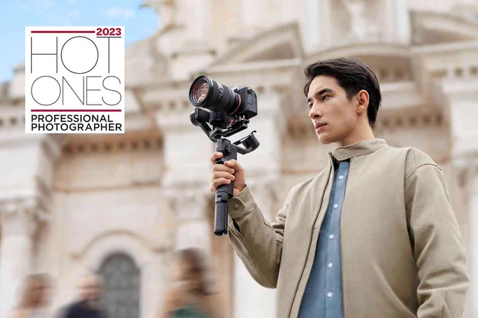 A man wearing a tan jacket a blue button up shirt is holding a camera in front of a building with text that says "2023 Hot Ones Professional Photographer."