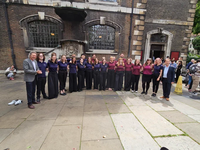 In a stone courtyard, the Genesis Sixteen cohort 12 singers stood together dressed in matching navy blue or claret t-shirts and smart black trousers. Either side of the group stands Harry Christophers and Eamonn Dougan. All are smiling at the camera.