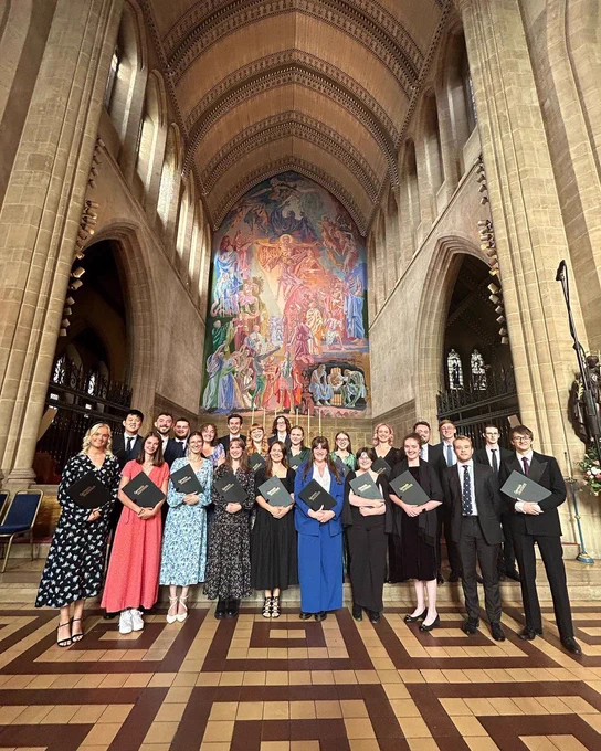 Inside a grand looking church, the Genesis Sixteen cohort 12 singers stood together, holding black scores and smiling at the camera. They are all dressed in smart suits or long dresses.