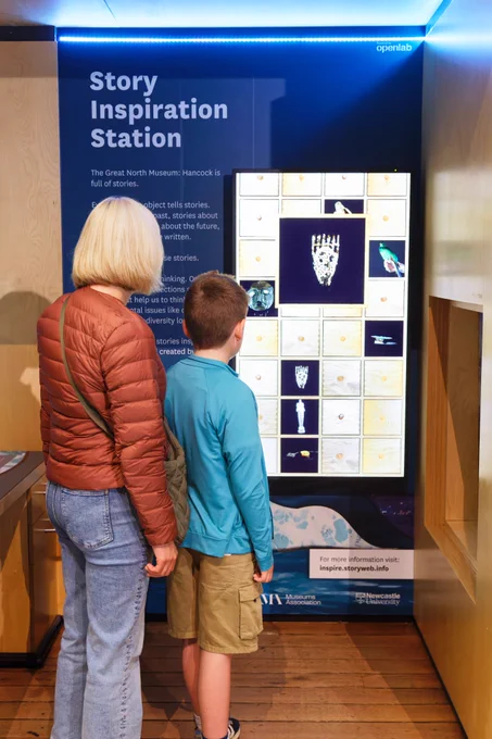 A woman and a child stand in front of the Story Inspiration Station. The Station is a large blue stand with a screen on it showing various images of objects in the museum. The text on the stand says: Story Inspiration Station. 