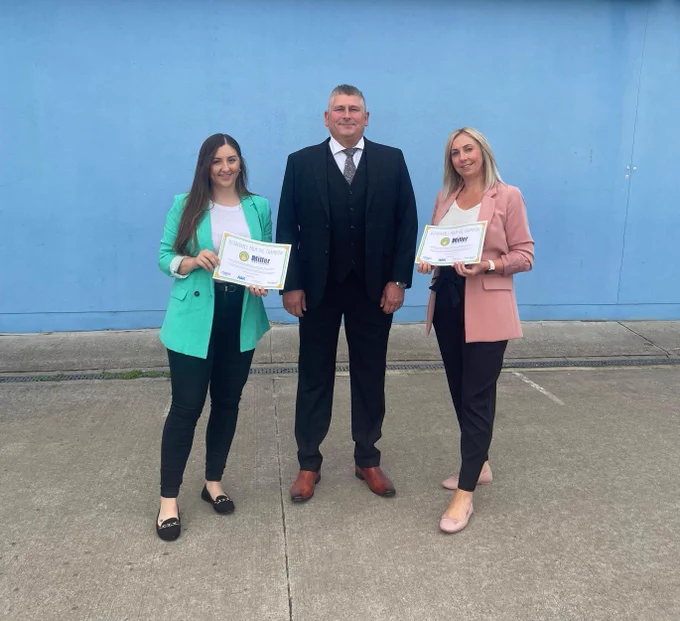 Two women stood either side of a man, holding certificates in front of The Deep's main entrance. 