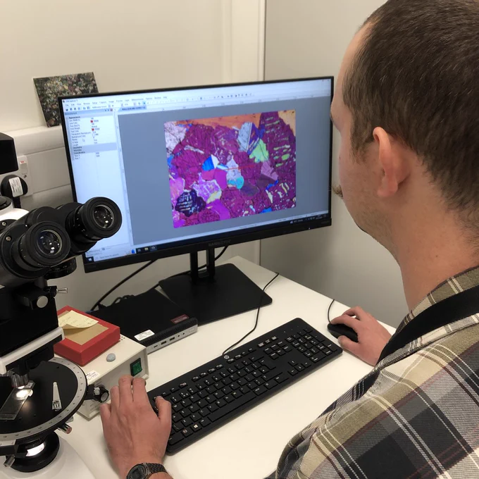 A man is sat at a computer attached to a polarising microscope. The screen is displaying a very pink thin section.