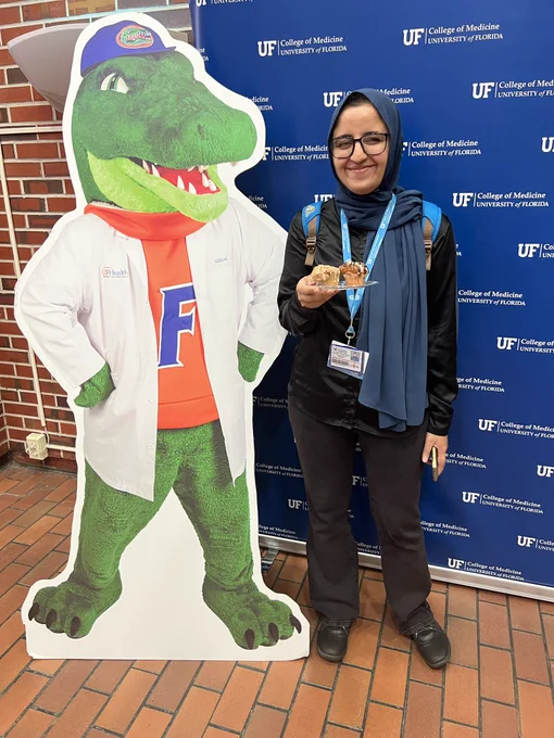 A person in black shoes, black pants, a black long-sleeve shirt and a blue headscarf smiling and holding a plate with two muffins next to a cutout of University of Florida mascot Albert E. Gator wearing a white coat.