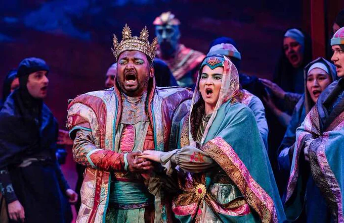 Two performers in colourful robes holding hands on stage, singing 