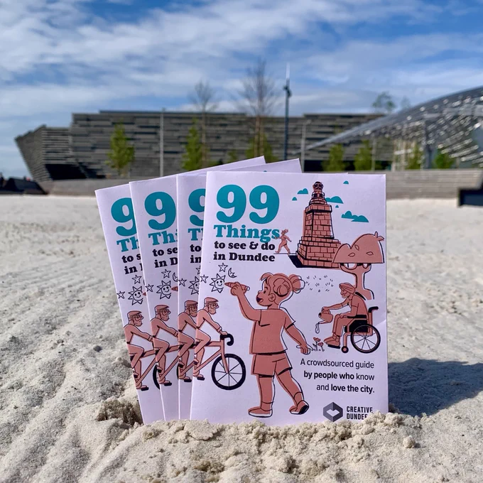 A fan of 4 copies of the 99 Things to See & Do in Dundee guide are propped up in the sand at Dundee's urban beach. The V&A Dundee can be seen in the background.