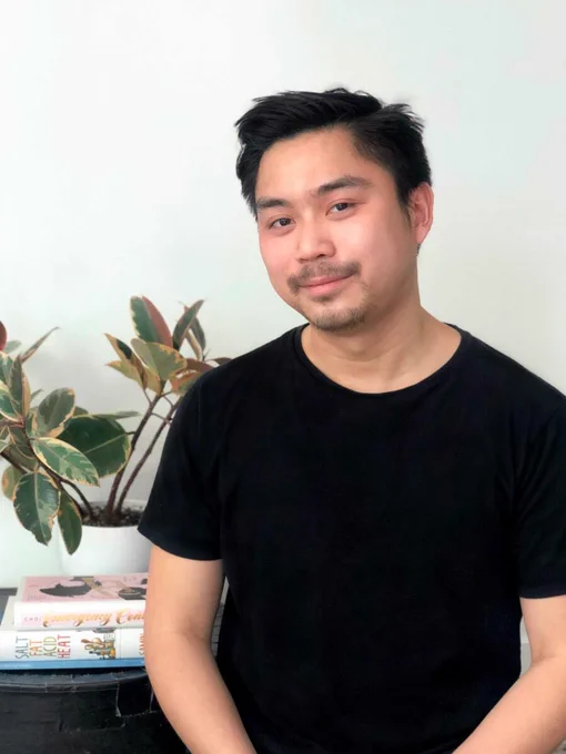 A male presenting person sits in front a white wall. They are smiling. There is a house plant in a white pot, and a book on a small table to their right side.