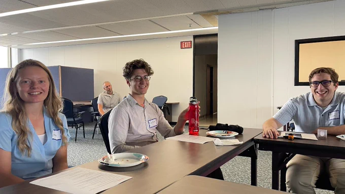 Three New York State Office of General Services student interns sit around a table and smile for the camera during the National Intern Day Forum.