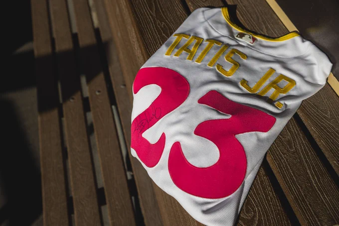 Photo of a City Connect jersey placed on a bench in the Padres dugout. The jersey is folded and you can see the back that reads “Tatis Jr.” in yellow and “23” in pink. Fernando’s signature is visible on the number 2.