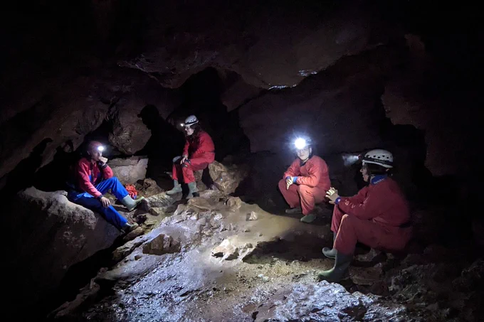 Four people in red caving suits, sitting in a cave chamber, surrounded by darkness. The space is lit by their headtorches. One person is looking directly out at us, their headtorch glaring. 