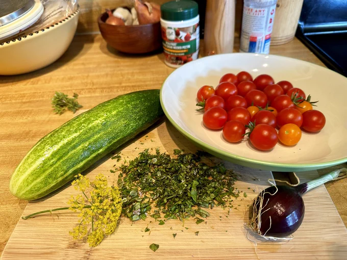 A cutting board holding a large cucumber, a bowl of cherry tomatoes, chopped herbs, a dill flower head, and a small red onion.